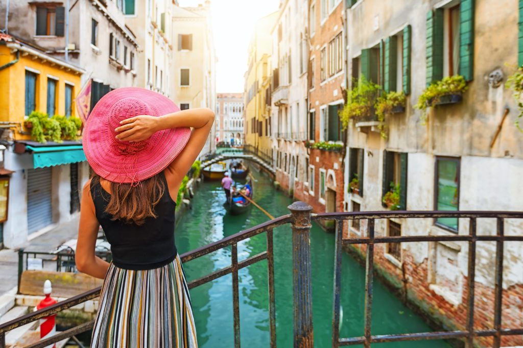 Frau mit rotem Sonnenhut schaut auf einen Kanal mit Gondel in Venedig, Italien Frau mit rotem Sonnenhut schaut auf einen Kanal mit Gondel in Venedig, Italien